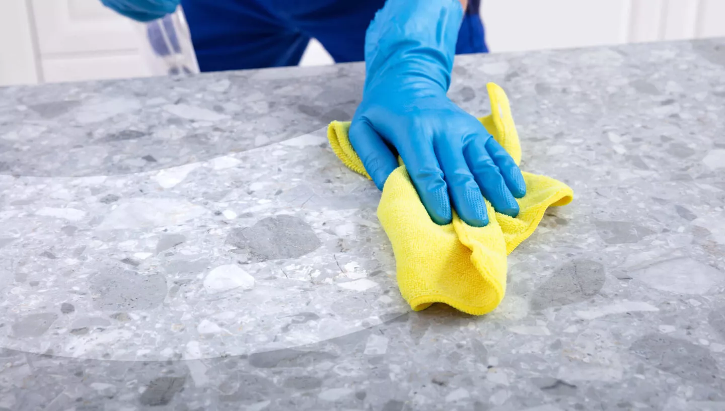 Eine Hand in blauem Handschuh wischt mit einem gelben Putzlappen über eine Keramikoberfläche. Die gewischt Oberfläche ist deutlich heller als die ungewischte.