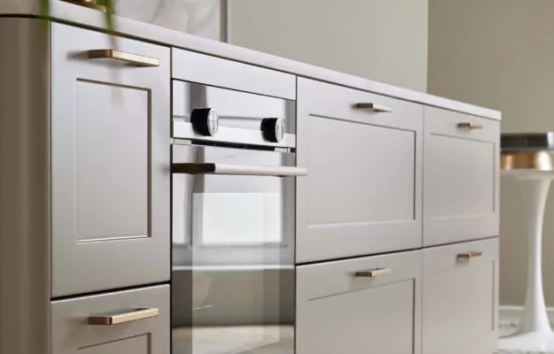 Nahaufnahme einer Kücheninsel mit graubeigen Rahmenfronten, goldfarbenen Möbelgriffen, heller Arbeitsplatte und ergonomisch integriertem Einbaubackofen.