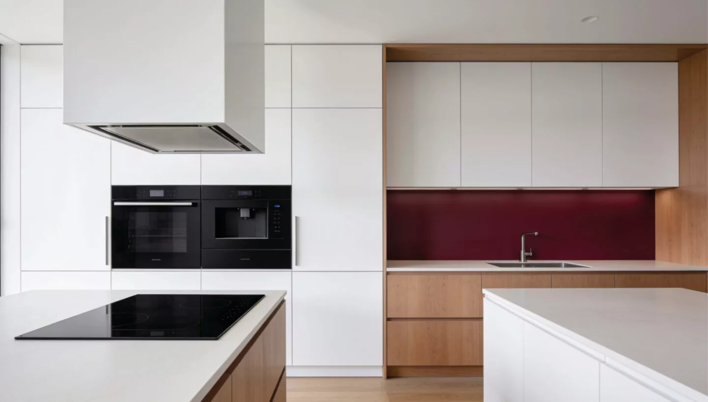 Minimalistische Designküche mit grifflosen weißen Hochschränken, warmen Holzfronten, bordeauxroter Rückwand, Einbaugeräten, Spüle und zwei Kücheninseln mit heller Arbeitsplatte und Induktionskochfeld.